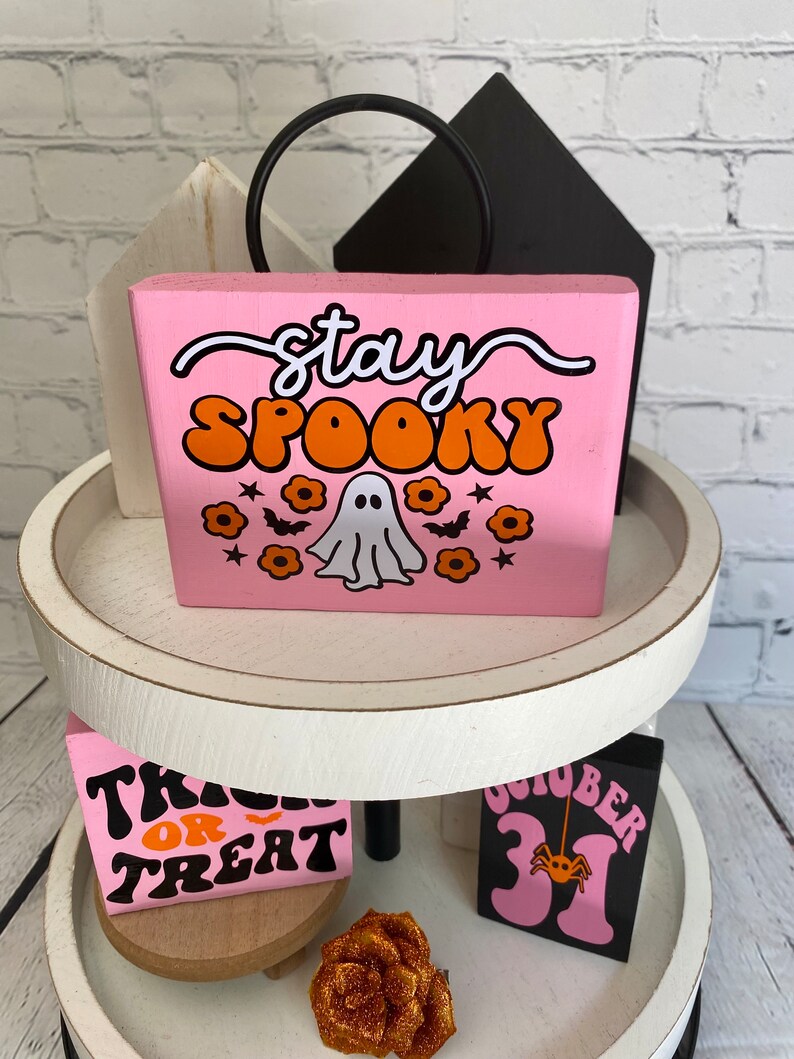 Puede incluir: Un letrero de madera rosa con el texto "stay spooky" en letras blancas y naranjas. Un fantasma blanco con ojos naranjas est&aacute; en el centro del letrero con flores naranjas y murci&eacute;lagos alrededor.