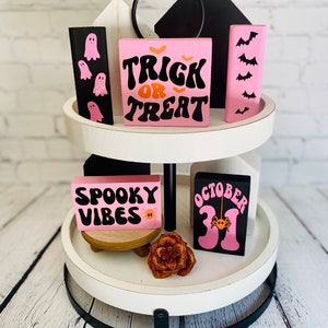 Op de afbeelding: Een wit etagerekastje met twee niveaus en een zwarte metalen basis. Het kastje is versierd met roze en zwarte houten blokken met Halloween-thema tekst en afbeeldingen, waaronder "Trick or Treat", "Spooky Vibes" en "October 31".