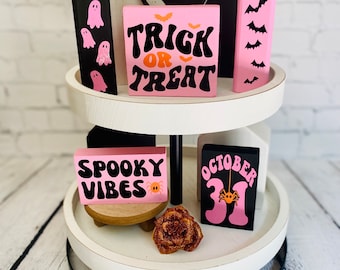 Op de afbeelding: Een wit etagerekastje met twee niveaus en een zwarte metalen basis. Het kastje is versierd met roze en zwarte houten blokken met Halloween-thema tekst en afbeeldingen, waaronder "Trick or Treat", "Spooky Vibes" en "October 31".
