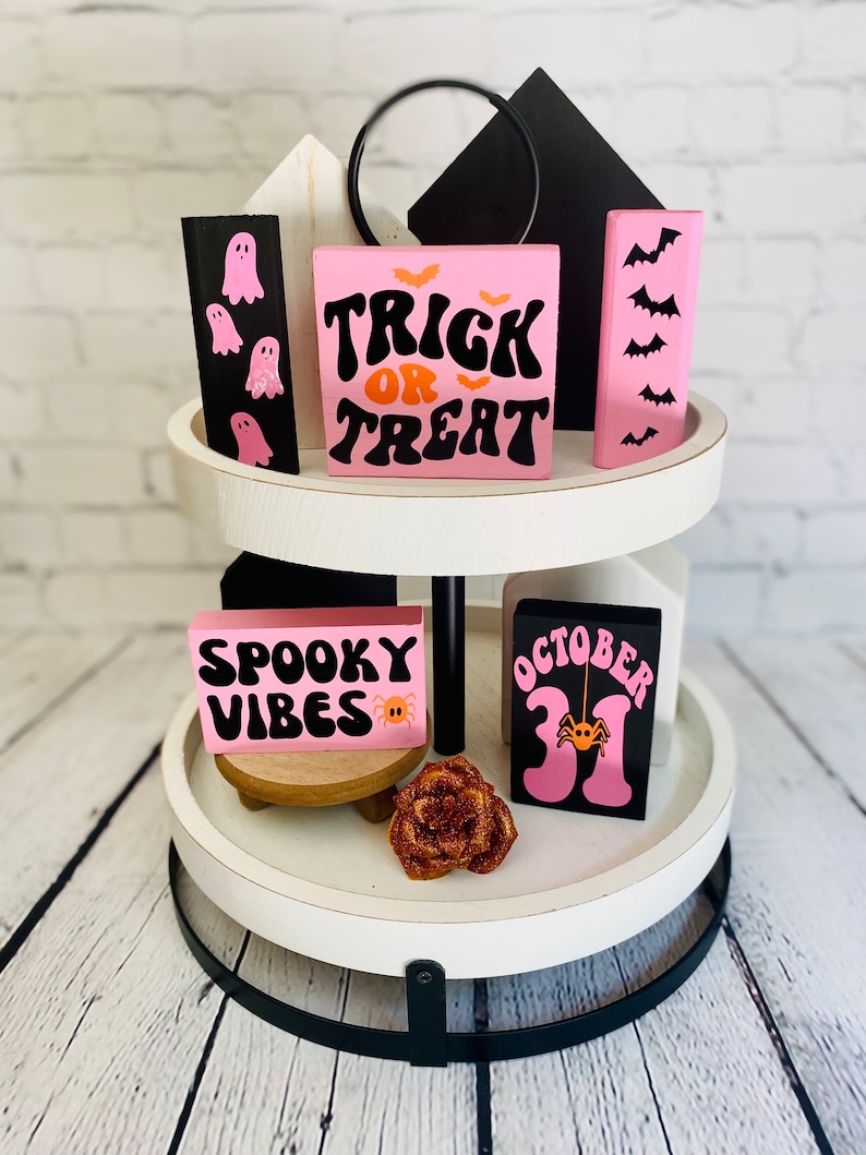 Op de afbeelding: Een wit etagerekastje met twee niveaus en een zwarte metalen basis. Het kastje is versierd met roze en zwarte houten blokken met Halloween-thema tekst en afbeeldingen, waaronder "Trick or Treat", "Spooky Vibes" en "October 31".