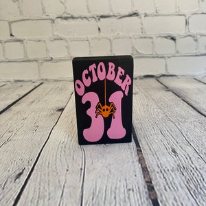 Op de afbeelding: Een zwarte houten blok met de woorden "OCTOBER 31" in roze letters. Een kleine oranje spin hangt aan de "1".