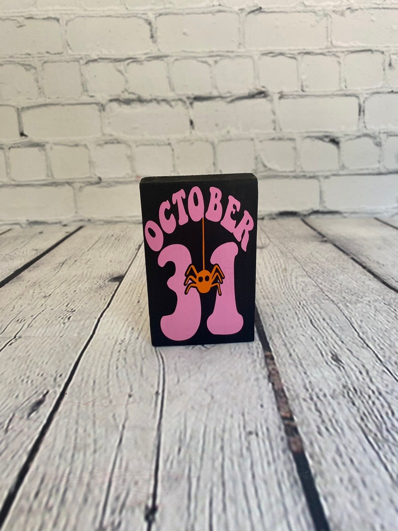Op de afbeelding: Een zwarte houten blok met de woorden "OCTOBER 31" in roze letters. Een kleine oranje spin hangt aan de "1".