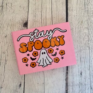 Puede incluir: Cuaderno rosa con el texto "stay spooky" en letras blancas y naranjas. Un fantasma blanco con ojos negros est&aacute; en el centro del cuaderno. Hay flores naranjas y murci&eacute;lagos negros alrededor del fantasma.