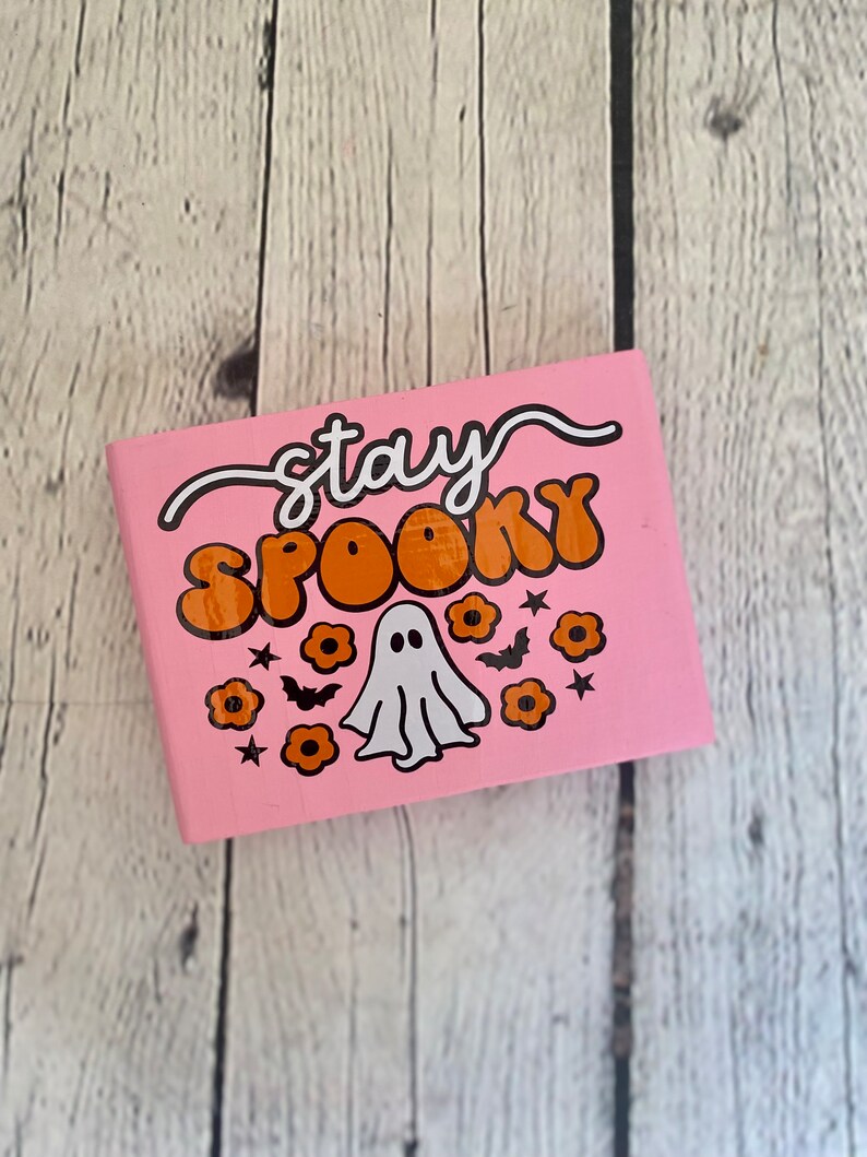 Puede incluir: Cuaderno rosa con el texto "stay spooky" en letras blancas y naranjas. Un fantasma blanco con ojos negros est&aacute; en el centro del cuaderno. Hay flores naranjas y murci&eacute;lagos negros alrededor del fantasma.