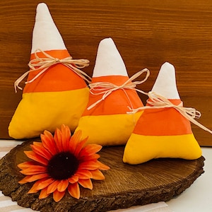 May include: Three fabric candy corn decorations in different sizes, with white, orange, and yellow stripes. The candy corn are sitting on a wooden slice with a single orange flower.