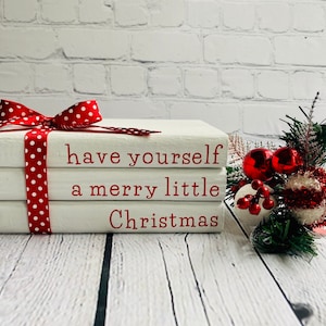 May include: A stack of three white books with red polka dot ribbon tied around the top book. The books are stacked on a white wooden surface. The top book has the text "have yourself a merry little Christmas" in red lettering.