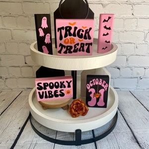 Op de afbeelding: Een wit etagerekastje met twee niveaus en een zwarte metalen basis. Het kastje is versierd met roze en zwarte houten bordjes met Halloween-thema's, waaronder "Trick or Treat", "Spooky Vibes" en "October 31".