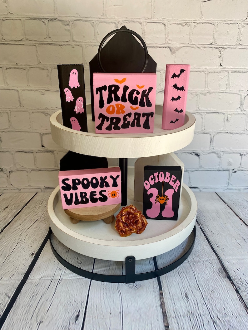 Op de afbeelding: Een wit etagerekastje met twee niveaus en een zwarte metalen basis. Het kastje is versierd met roze en zwarte houten bordjes met Halloween-thema's, waaronder "Trick or Treat", "Spooky Vibes" en "October 31".