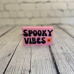 Op de afbeelding: Roze houten blok met de tekst "Spooky Vibes" in zwarte letters en een kleine oranje spin afbeelding.