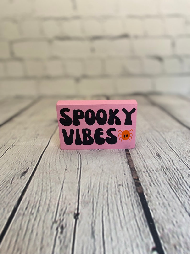 Op de afbeelding: Roze houten blok met de tekst "Spooky Vibes" in zwarte letters en een kleine oranje spin afbeelding.