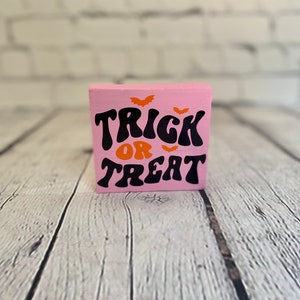 Op de afbeelding: Een roze houten blok met de woorden "Trick or Treat" in zwarte en oranje letters. Er zijn drie oranje vleermuizen boven de tekst.