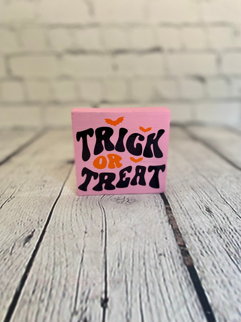 Op de afbeelding: Een roze houten blok met de woorden "Trick or Treat" in zwarte en oranje letters. Er zijn drie oranje vleermuizen boven de tekst.