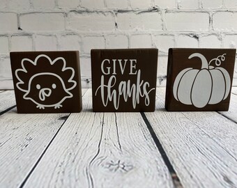 May include: Three brown wooden blocks with white painted designs. The first block has a turkey, the second block says "GIVE THANKS", and the third block has a pumpkin.