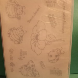 May include: A sheet of clear cling stamps featuring various images of ladybugs, flowers, and inspirational phrases in French, including "Bonne chance!" (Good luck!), "Je pense a toi" (I'm thinking of you), "Ouvre tes ailes et vole!" (Open your wings and fly!), and "Tu me portes bonheur" (You bring me luck).