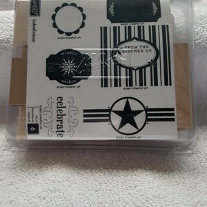 May include: A set of six clear plastic-packaged rubber stamps with black ink. The stamps feature various designs, including a circle, a star, a banner with the word "celebrate", a banner with the words "winter wishes", and a banner with the words "from the kitchen of".
