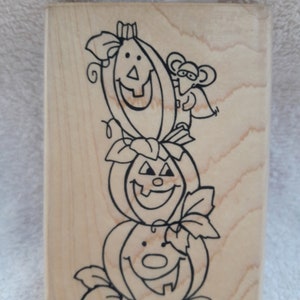 May include: A wooden stamp with a black and white illustration of three pumpkins with jack-o-lantern faces and a mouse climbing on top of the pumpkins.