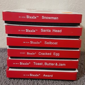 May include: A stack of six red and black Sizzix die-cutting machine dies. The dies are labeled with the following names: Snowman, Santa Head, Sailboat, Cracked Egg, Toast, Butter & Jam, and Award. Each die has a corresponding number: 38-0235, 38-0233, 38-0124, 38-0257, 38-0914, and 38-0139.
