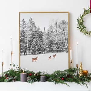 Könnte beinhalten: Ein gerahmter Winterlandschaftsdruck mit Hirschen in einem schneebedeckten Feld, mit hohen immergrünen Bäumen im Hintergrund. Die Szene ist mit Kerzen, einer Girlande und einem Kranz dekoriert und schafft eine festliche Atmosphäre.