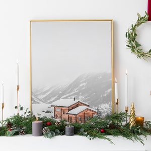 K&ouml;nnte beinhalten: Ein gerahmter Druck einer schneebedeckten Berglandschaft mit einer Holzh&uuml;tte. Der Kaminsims ist mit Gr&uuml;n, Kerzen und einem goldenen Weihnachtsbaum geschm&uuml;ckt. Eine Girlande h&auml;ngt dar&uuml;ber und schafft eine festliche Atmosph&auml;re.