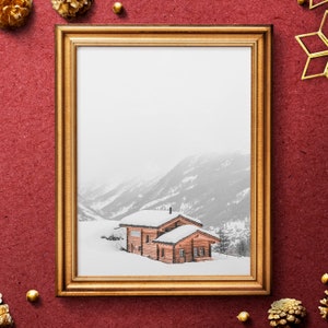K&ouml;nnte beinhalten: Ein gerahmter Druck mit einem schneebedeckten Holzhaus in einem Bergtal. Das Haus hat ein schneebedecktes Dach und ist von schneebedeckten B&auml;umen umgeben. Der Rahmen ist goldfarben und der Hintergrund ist strukturiertes Rot. Goldene Tannenzapfen und ein Stern schm&uuml;cken die Ecken.