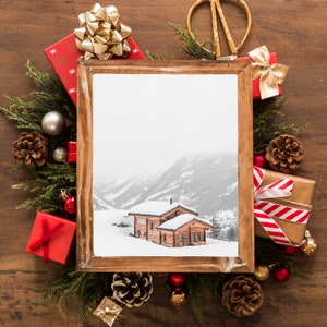K&ouml;nnte beinhalten: Ein holzgerahmtes Bild eines schneebedeckten Ferienhauses in einer Winterlandschaft, umgeben von Weihnachtsdekorationen. Rote und braune Geschenkverpackungen mit goldenen und roten Schleifen, Tannenzapfen und immergr&uuml;ne Zweige sind auf einer Holzoberfl&auml;che angeordnet.