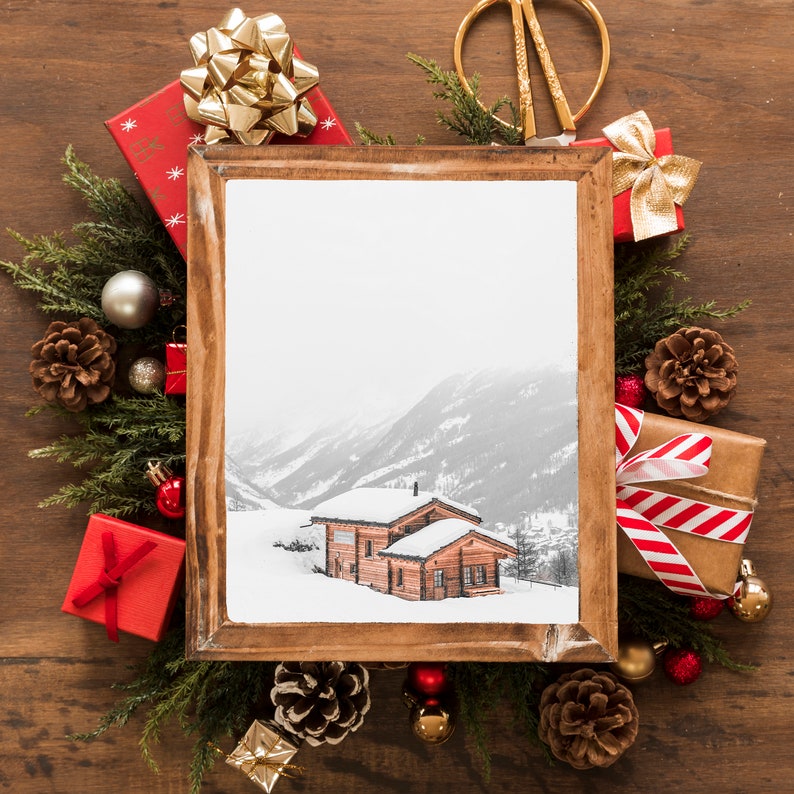 K&ouml;nnte beinhalten: Ein holzgerahmtes Bild eines schneebedeckten Ferienhauses in einer Winterlandschaft, umgeben von Weihnachtsdekorationen. Rote und braune Geschenkverpackungen mit goldenen und roten Schleifen, Tannenzapfen und immergr&uuml;ne Zweige sind auf einer Holzoberfl&auml;che angeordnet.