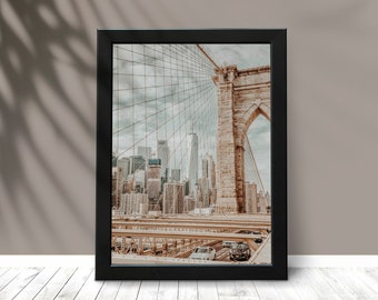 May include: Framed photograph of the Brooklyn Bridge in New York City, featuring the bridge's architecture and the city skyline. The image is in a black frame and is displayed on a white wooden surface.