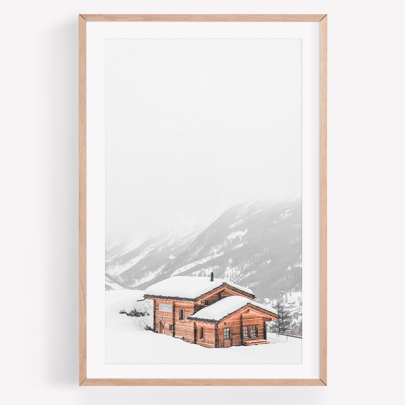 K&ouml;nnte beinhalten: Gerahmter Druck mit einem schneebedeckten Holzchalet in einer Berglandschaft. Das Chalet hat ein schneebedecktes Dach und ist von schneebedecktem Gel&auml;nde umgeben. Die Berge sind im Hintergrund und der Himmel ist bedeckt.