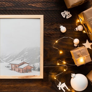 K&ouml;nnte beinhalten: Ein gerahmtes Bild einer verschneiten Berglandschaft mit Holzh&uuml;tten. Das Bild liegt auf einer dunklen Holzoberfl&auml;che mit verpackten Geschenken, Lichterketten und Weihnachtsschmuck.