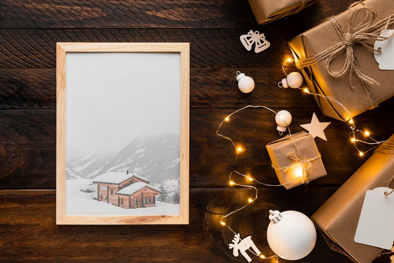 K&ouml;nnte beinhalten: Ein gerahmtes Bild einer verschneiten Berglandschaft mit Holzh&uuml;tten. Das Bild liegt auf einer dunklen Holzoberfl&auml;che mit verpackten Geschenken, Lichterketten und Weihnachtsschmuck.