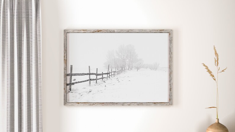 以下が含まれることがあります： 雪景色を捉えた、額装された白黒写真。 雪に覆われた野原に沿って木製の柵が伸び、背景には木々が見えます。 フレームは明るく風化した木材です。 全体的なテーマは冬の風景です。