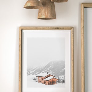 K&ouml;nnte beinhalten: Gerahmter Druck einer schneebedeckten Berglandschaft mit einer Holzh&uuml;tte. Der Druck befindet sich in einem hellen Holzrahmen, und das Bild spiegelt sich auf der darunter liegenden Oberfl&auml;che. Drei Pendelleuchten h&auml;ngen &uuml;ber dem Rahmen.