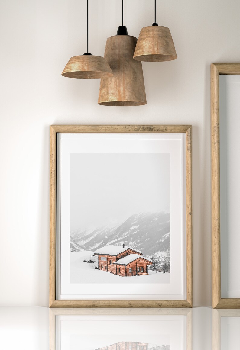 K&ouml;nnte beinhalten: Gerahmter Druck einer schneebedeckten Berglandschaft mit einer Holzh&uuml;tte. Der Druck befindet sich in einem hellen Holzrahmen, und das Bild spiegelt sich auf der darunter liegenden Oberfl&auml;che. Drei Pendelleuchten h&auml;ngen &uuml;ber dem Rahmen.