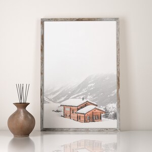K&ouml;nnte beinhalten: Ein gerahmter Druck einer verschneiten Berglandschaft mit zwei Holzh&uuml;tten. Die H&uuml;tten haben schneebedeckte D&auml;cher und liegen in einem Tal. Links vom Rahmen befindet sich eine braune Vase mit schwarzen St&auml;ben.