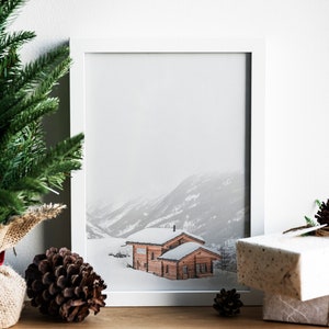 K&ouml;nnte beinhalten: Ein gerahmter Druck einer verschneiten Berglandschaft mit zwei Holzh&uuml;tten. Der Druck in einem wei&szlig;en Rahmen ist von Weihnachtsdekorationen umgeben, darunter ein Tannenbaum, Tannenzapfen und verpackte Geschenke.