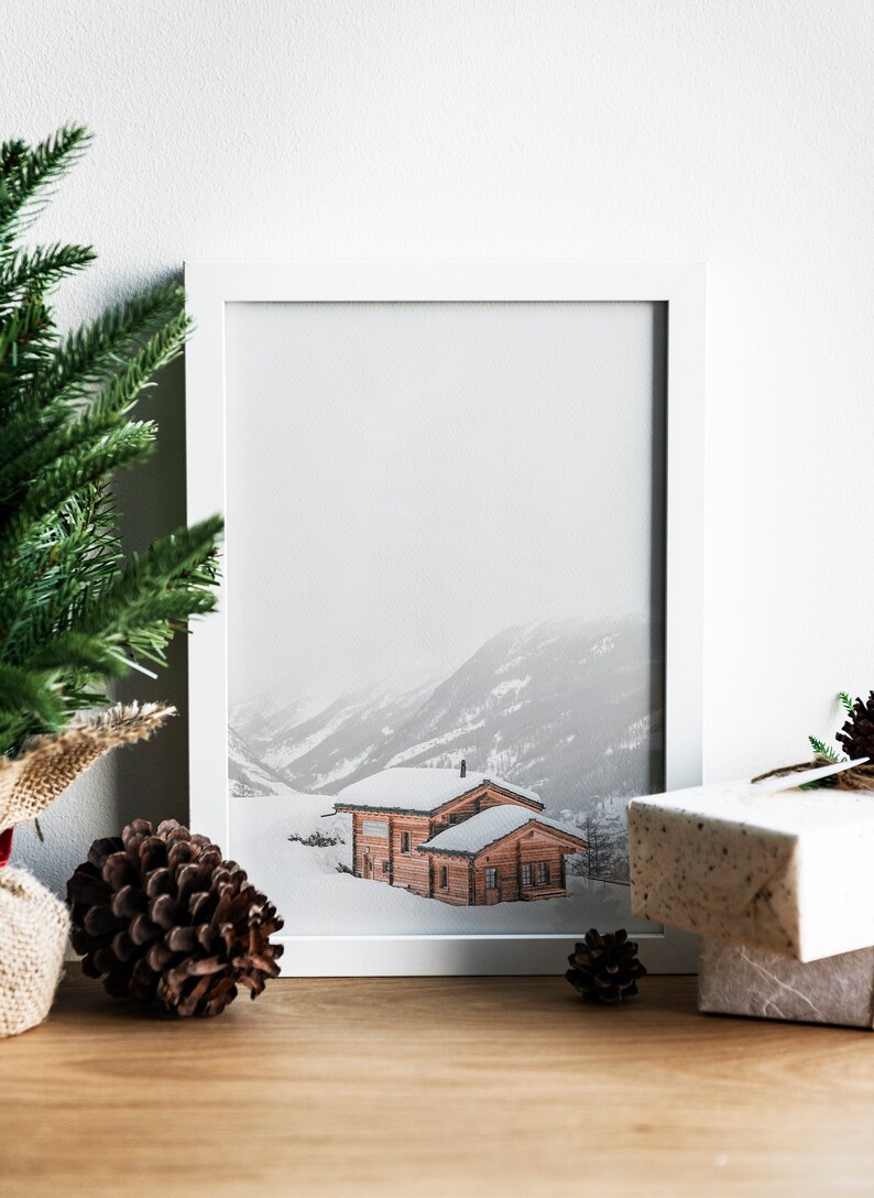 K&ouml;nnte beinhalten: Ein gerahmter Druck einer verschneiten Berglandschaft mit zwei Holzh&uuml;tten. Der Druck in einem wei&szlig;en Rahmen ist von Weihnachtsdekorationen umgeben, darunter ein Tannenbaum, Tannenzapfen und verpackte Geschenke.