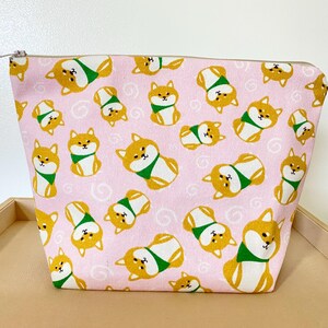 May include: Pink zippered pouch with a repeating pattern of Shiba Inu dogs wearing green scarves. The dogs are yellow and white with black eyes and noses. The pouch has a silver zipper.
