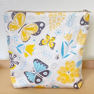 May include: A white zippered pouch with a floral and butterfly design. The butterflies are yellow, blue, and gray. The flowers are yellow and white.