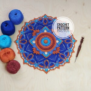 Op de afbeelding: Een gehaakt mandala-patroon in tinten blauw, oranje en turquoise. Het patroon is rond en heeft een herhalend ontwerp van bloemen en bladeren. Het patroon is omgeven door een rand van gehaakte steken. De tekst "CROCHET PATTERN ENG/RUS" staat in een cirkel aan de rechterkant van de afbeelding.
