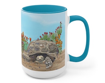 May include: A white ceramic mug with a turquoise rim and handle. The mug features a colorful illustration of a desert tortoise in a desert landscape with cacti and flowers.
