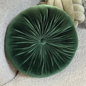 May include: A round, dark green velvet pillow with a button in the center. The pillow has a tufted design.