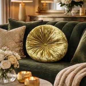 May include: A round, gold velvet pillow with a button center sits on a dark green sofa. The pillow has a radial design and is surrounded by other pillows and decorative items, creating a luxurious living room setting.