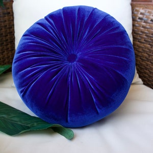 May include: A round, blue velvet pillow with a button tufting design. The pillow is soft and plush, and would be a great addition to any living room or bedroom.