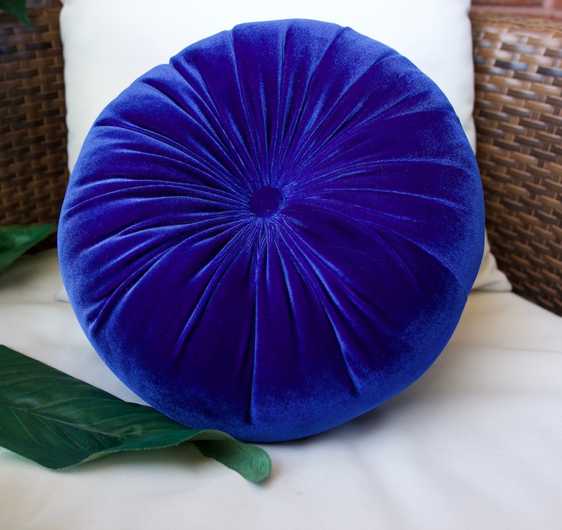May include: A round, blue velvet pillow with a button tufting design. The pillow is soft and plush, and would be a great addition to any living room or bedroom.