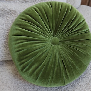 May include: A round, olive green velvet pillow with a button center. The pillow has a pleated design radiating from the center button. The pillow is on a light-colored, textured surface.