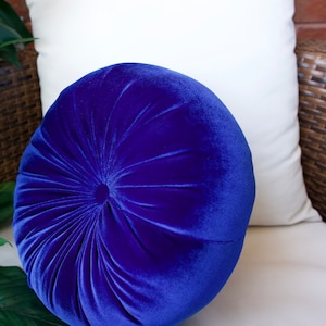 May include: A round, blue velvet pillow with a button tufting design. The pillow is sitting on a white couch.