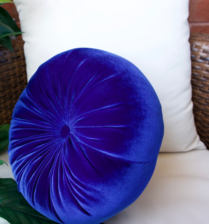 May include: A round, blue velvet pillow with a button tufting design. The pillow is sitting on a white couch.