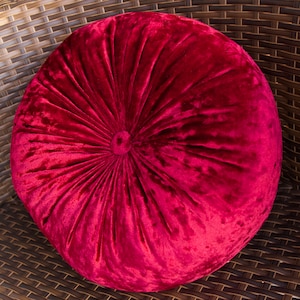 May include: A round, plush cushion in a deep red hue. The velvet fabric is gathered towards the center, creating a textured, radial pattern. The cushion is placed on a woven, brown surface.