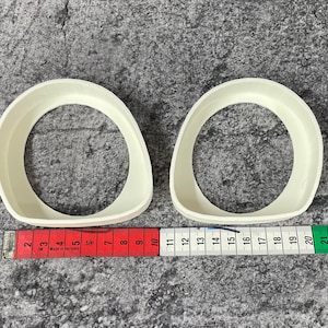 Op de afbeelding: Twee witte plastic ringen, elk met een diameter van ongeveer 25 cm. De ringen liggen op een grijs, getextureerd oppervlak met een rood en groen meetlint.