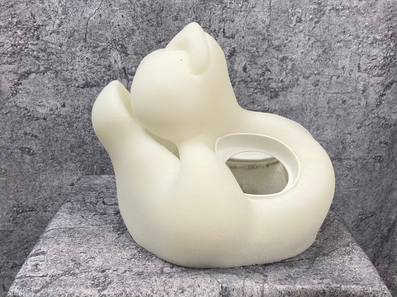 K&ouml;nnte beinhalten: Wei&szlig;er Keramik-Pflanztopf in Form einer zusammengerollten Katze mit einer runden &Ouml;ffnung f&uuml;r eine Pflanze.